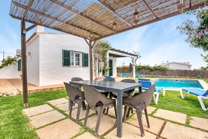Une terrasse couverte avec une table et des chaises, une piscine et un bâtiment blanc aux volets verts.