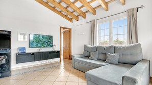 Een moderne woonkamer met een grijze bank, houten balkenplafond, een televisie en een groot raam met gordijnen.