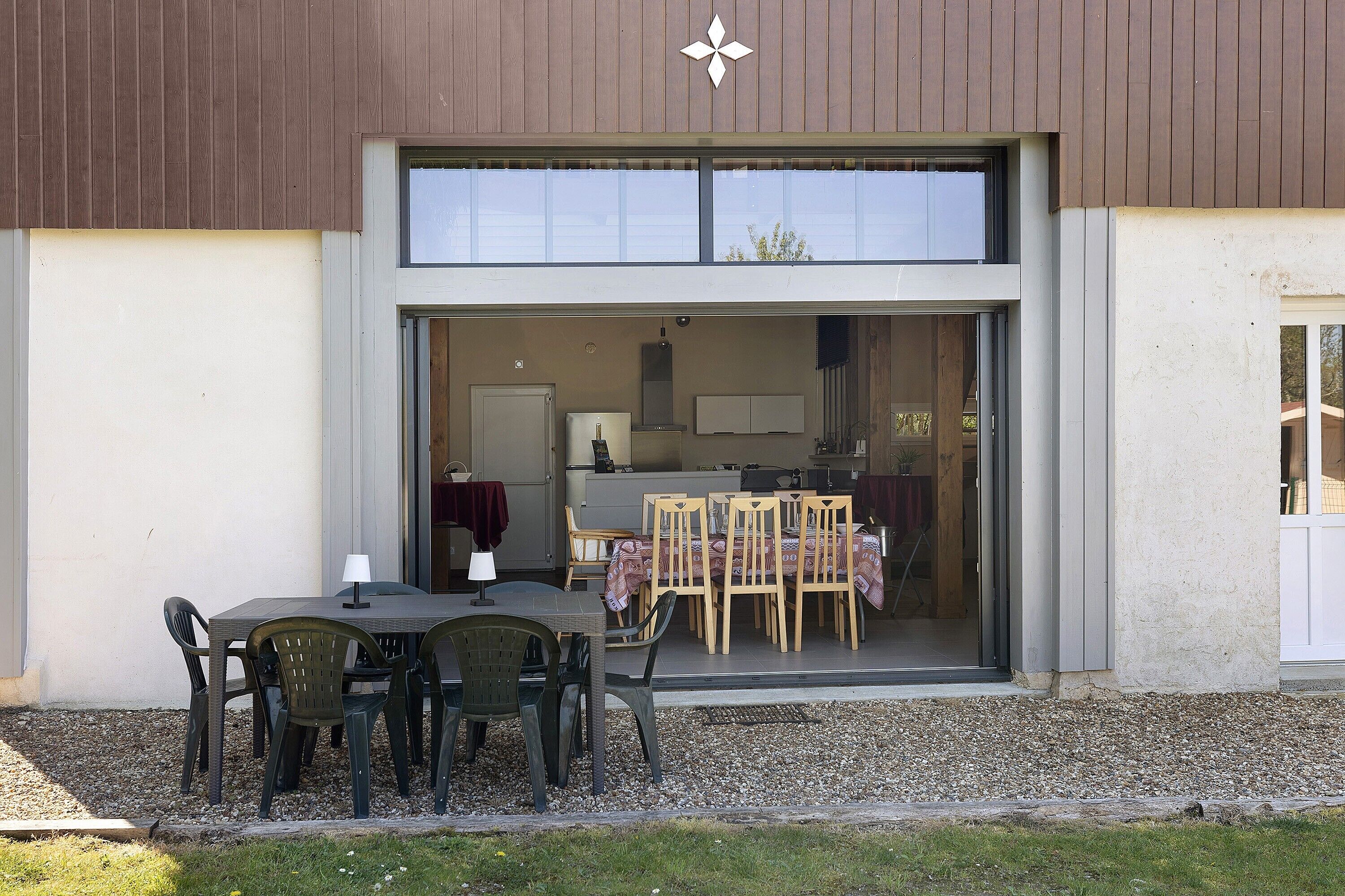 Een modern gebouw met een buiten eetgedeelte, inclusief een tafel en stoelen, een keuken zichtbaar door de glazen deur en een grindpad.