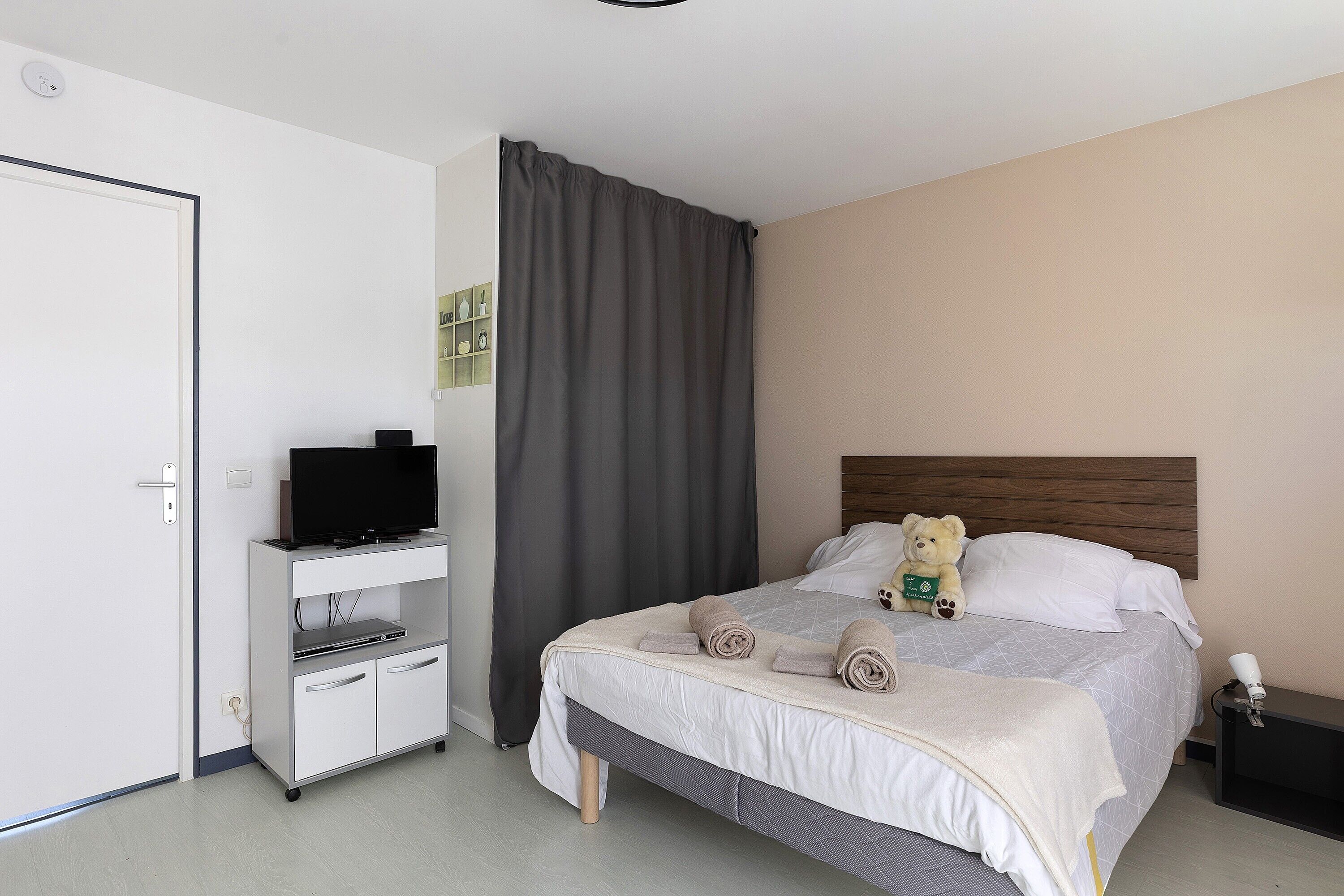 Een net opgemaakt bed met kussens en een teddybeer, een televisie op een standaard en een nachtkastje met een lamp.