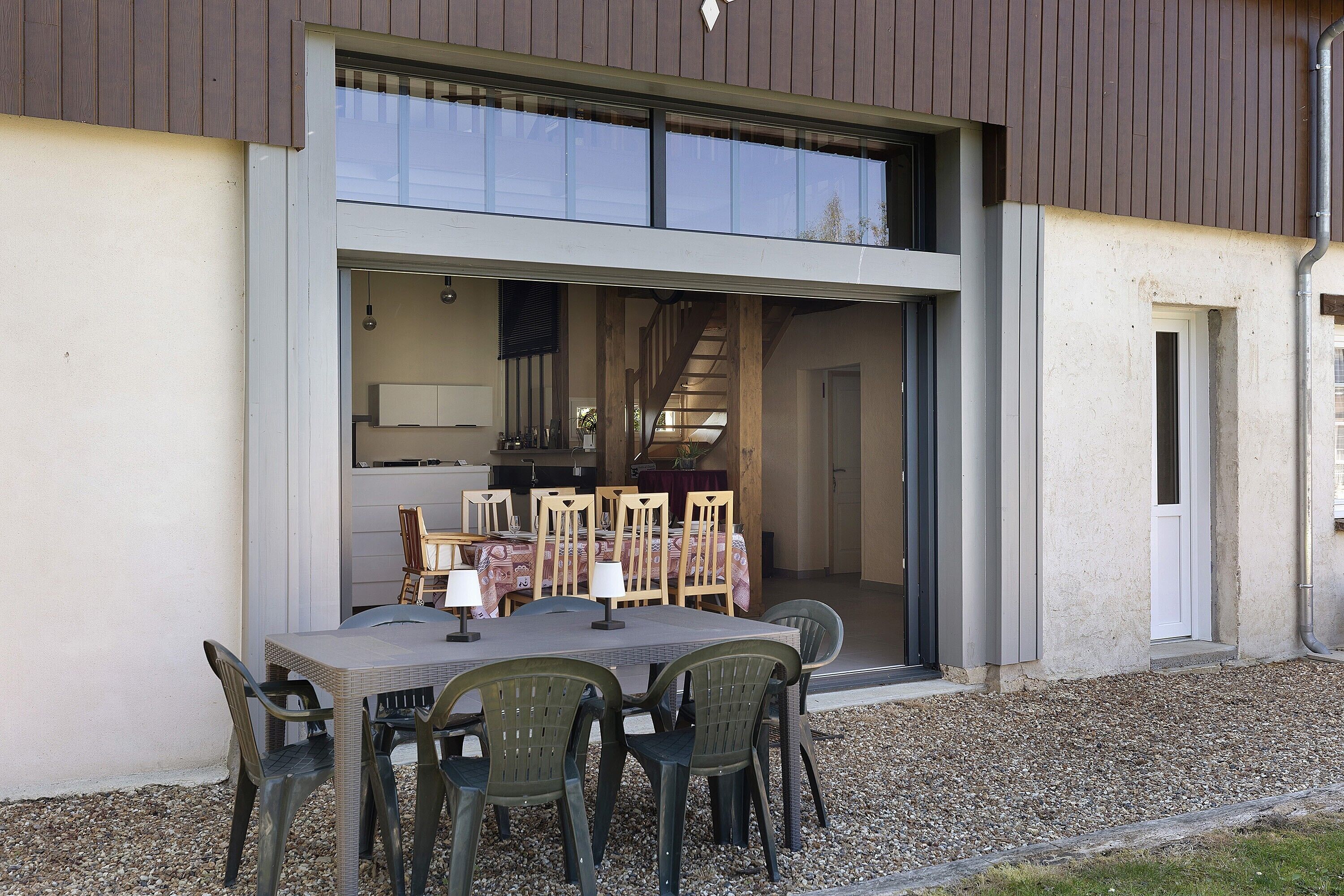 A modern building with a dining area featuring a table and chairs, a kitchen visible through an open door, and a gravel ground.