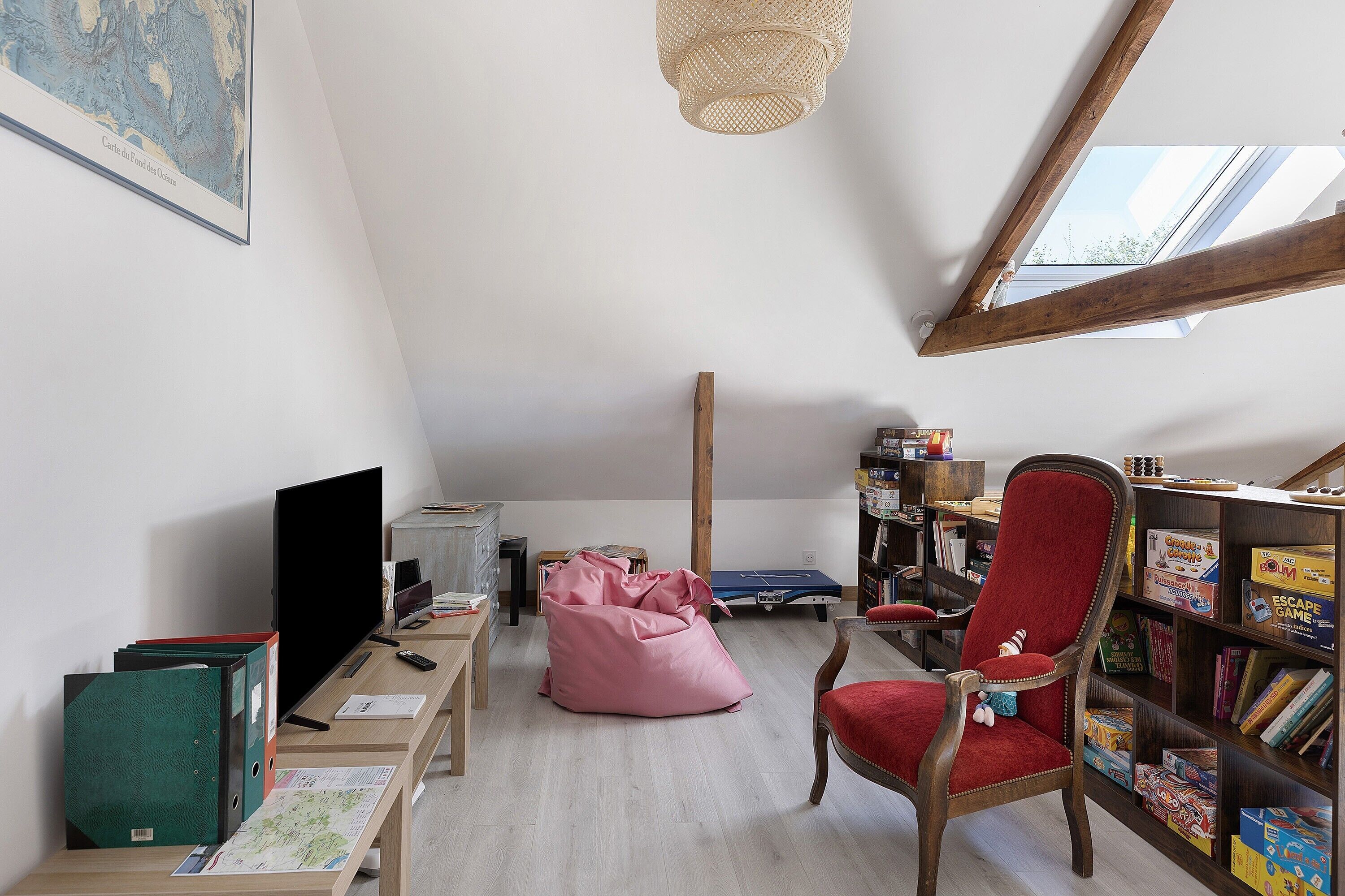 Een loftachtige kamer met schuine wanden, een krukkonenbank, een rode fauteuil, een boekenkast en een bureau met een computermonitor.