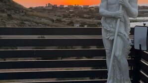 Een standbeeld van een figuur met een drietand, geplaatst op een witte bank met uitzicht op een zonsondergang en gebouwen op de achtergrond.