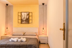 Un dormitorio con cama, almohadas, una mesita de noche, un cuadro en la pared y dos lámparas colgantes.