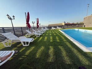 Zona de piscina con sillas reclinables blancas y sombrillas rojas, cielo azul despejado y vista a montañas distantes.