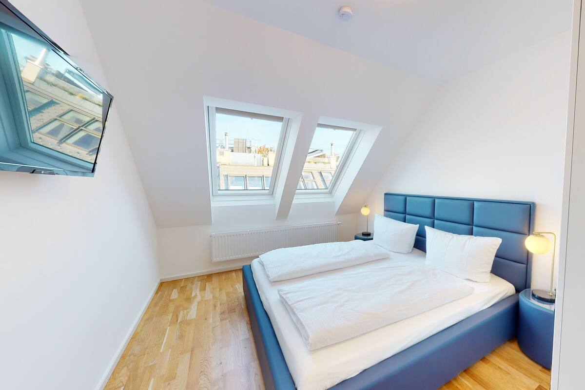 Une chambre moderne avec un grand lit, deux fenêtres, un ventilateur de plafond et une table de chevet avec une lampe.