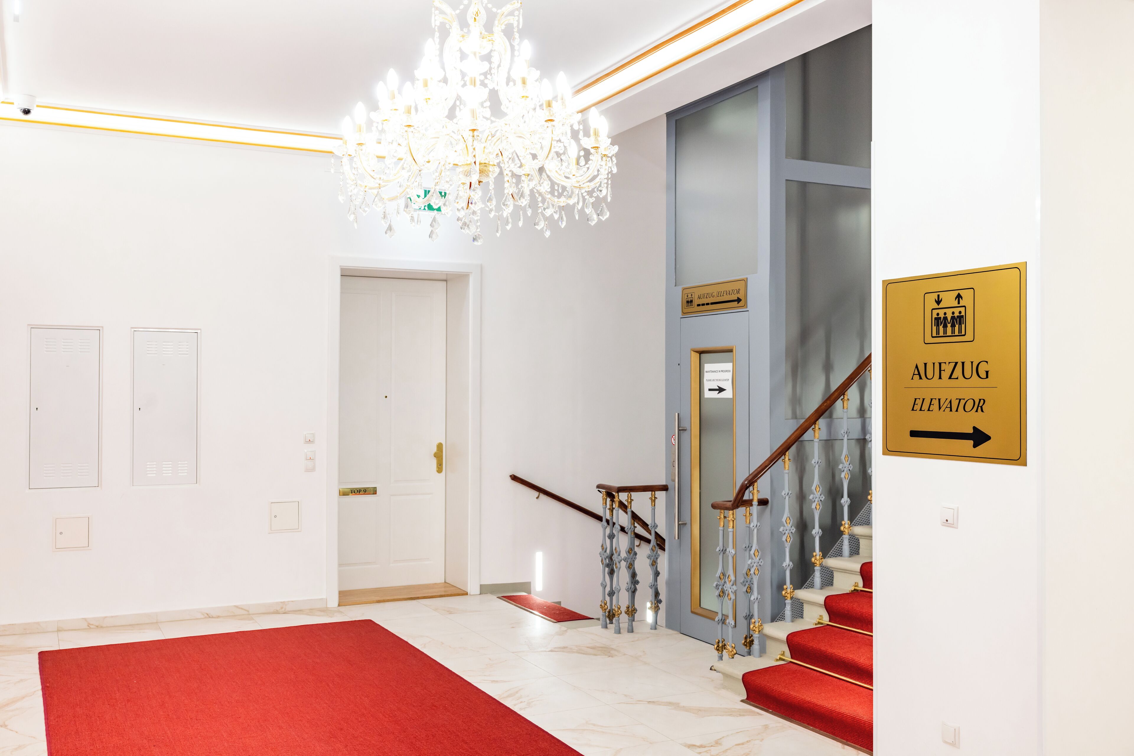A hotel lobby with a red carpet, a chandelier, a staircase with red steps, and an elevator sign.