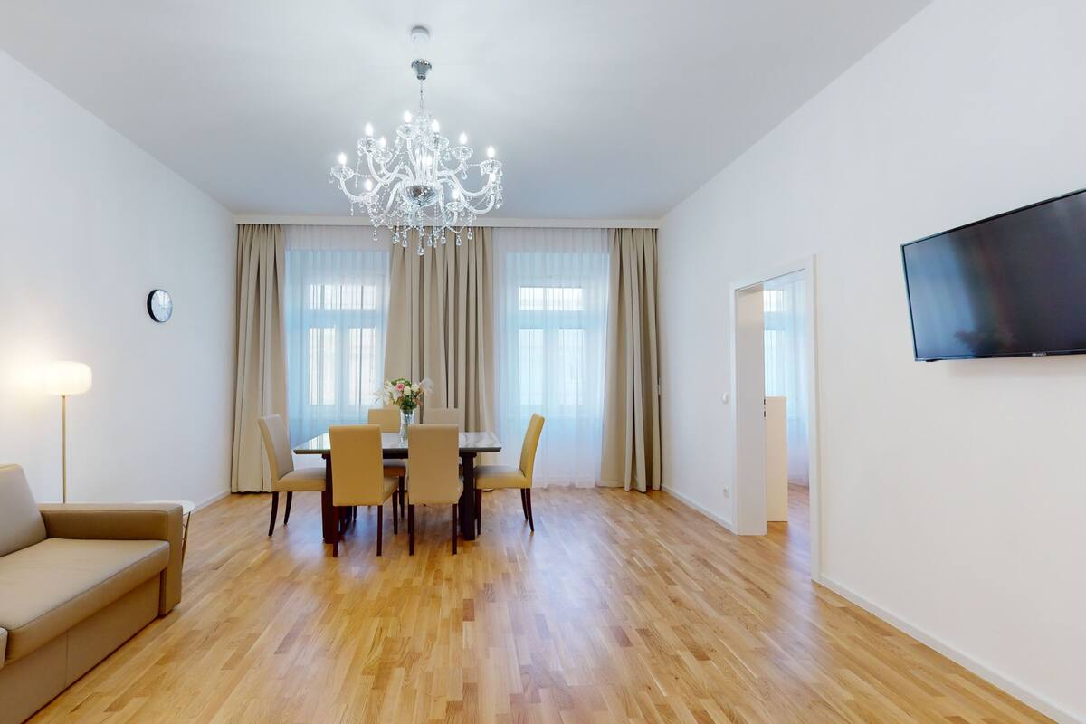 Un salon moderne avec un canapé beige, une table à manger avec des chaises, un lustre et un téléviseur à écran plat fixé au mur.