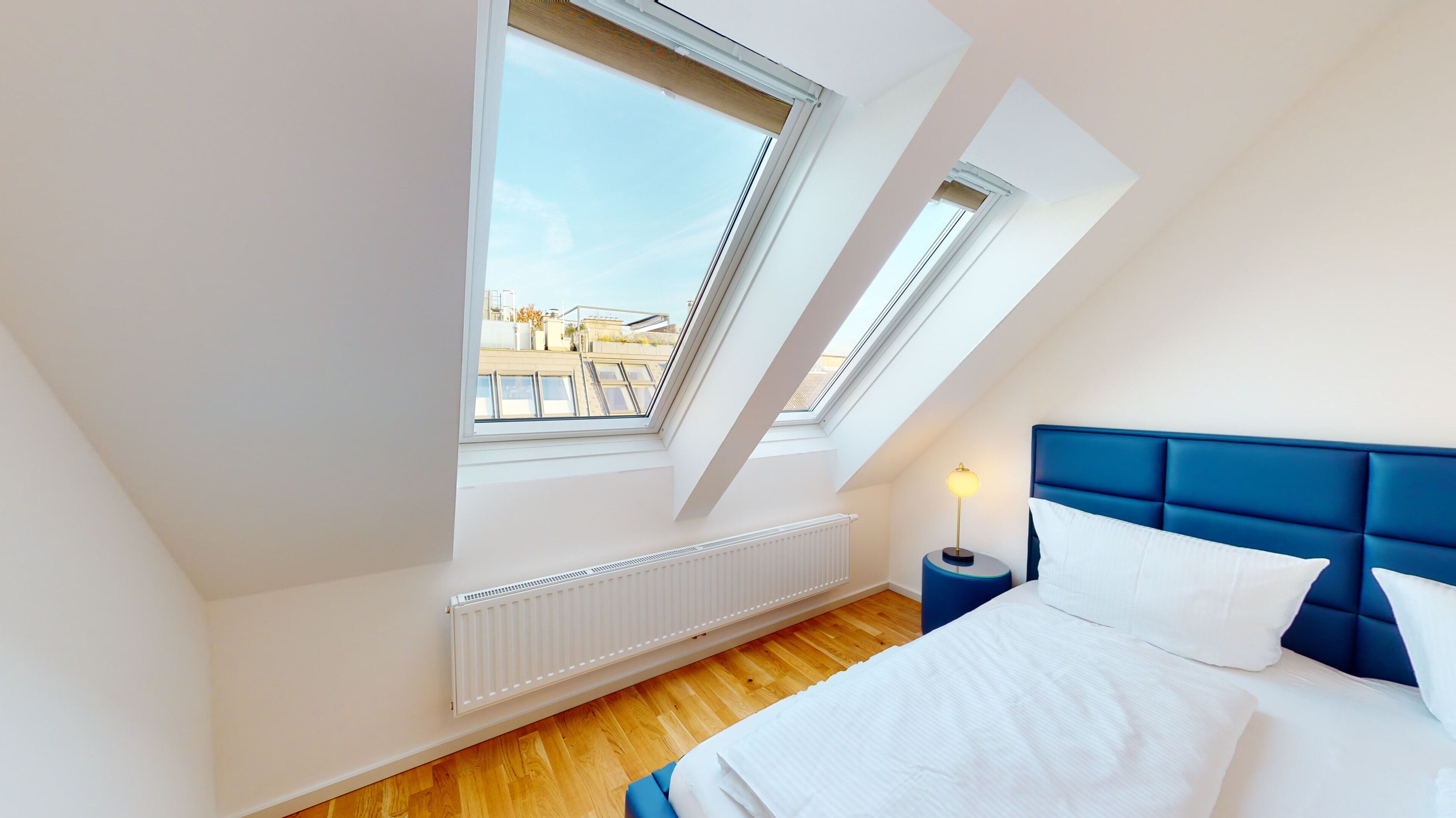 Un dormitorio con techo inclinado, ventanas de tragaluces, radiador, cama con ropa de cama blanca, cabecera azul y mesita de noche con lámpara.