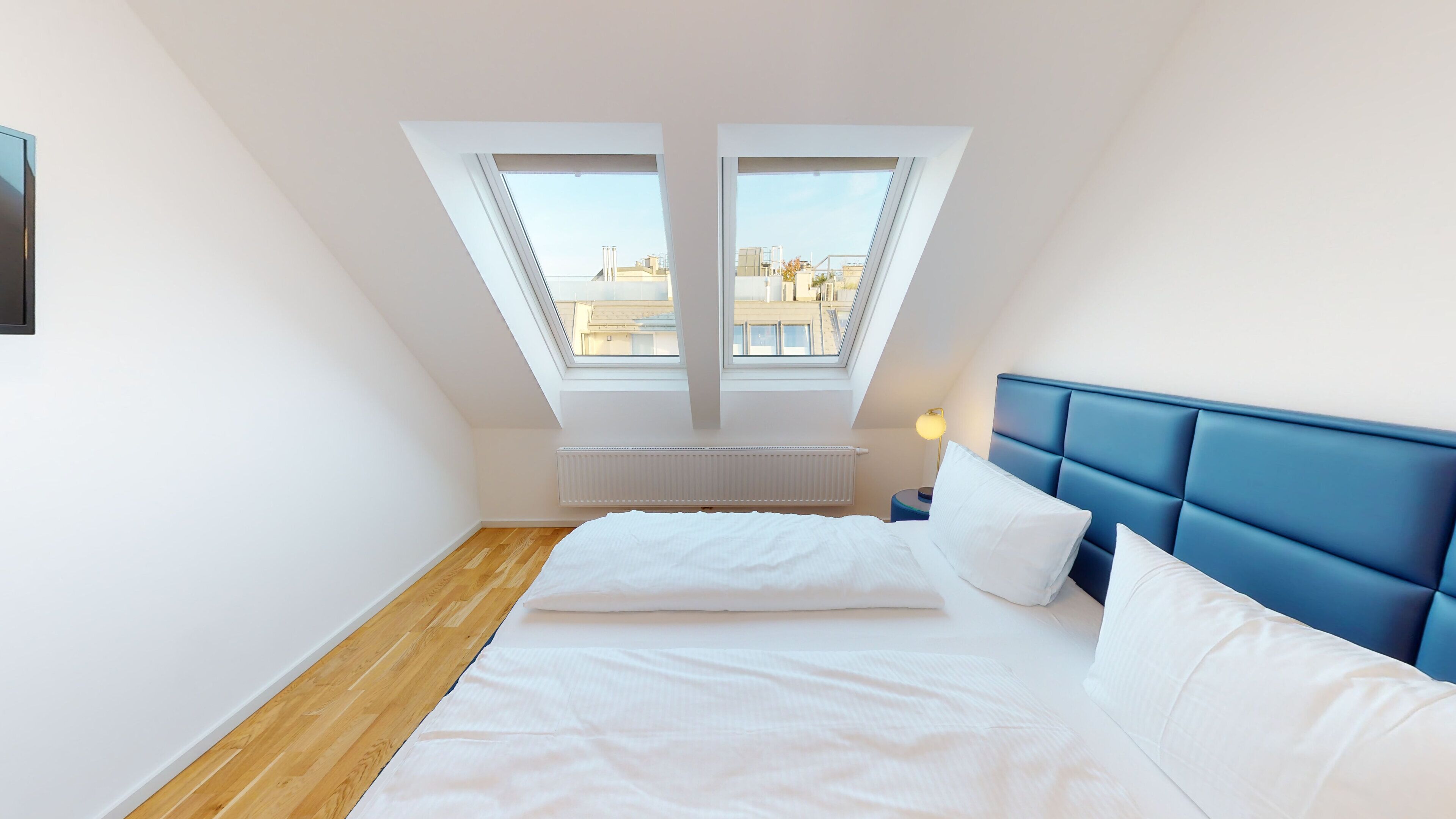 Une chambre avec un plafond en pente, deux fenêtres de toit, un lit avec une literie blanche, une tête de lit bleue et une table de chevet avec une lampe.
