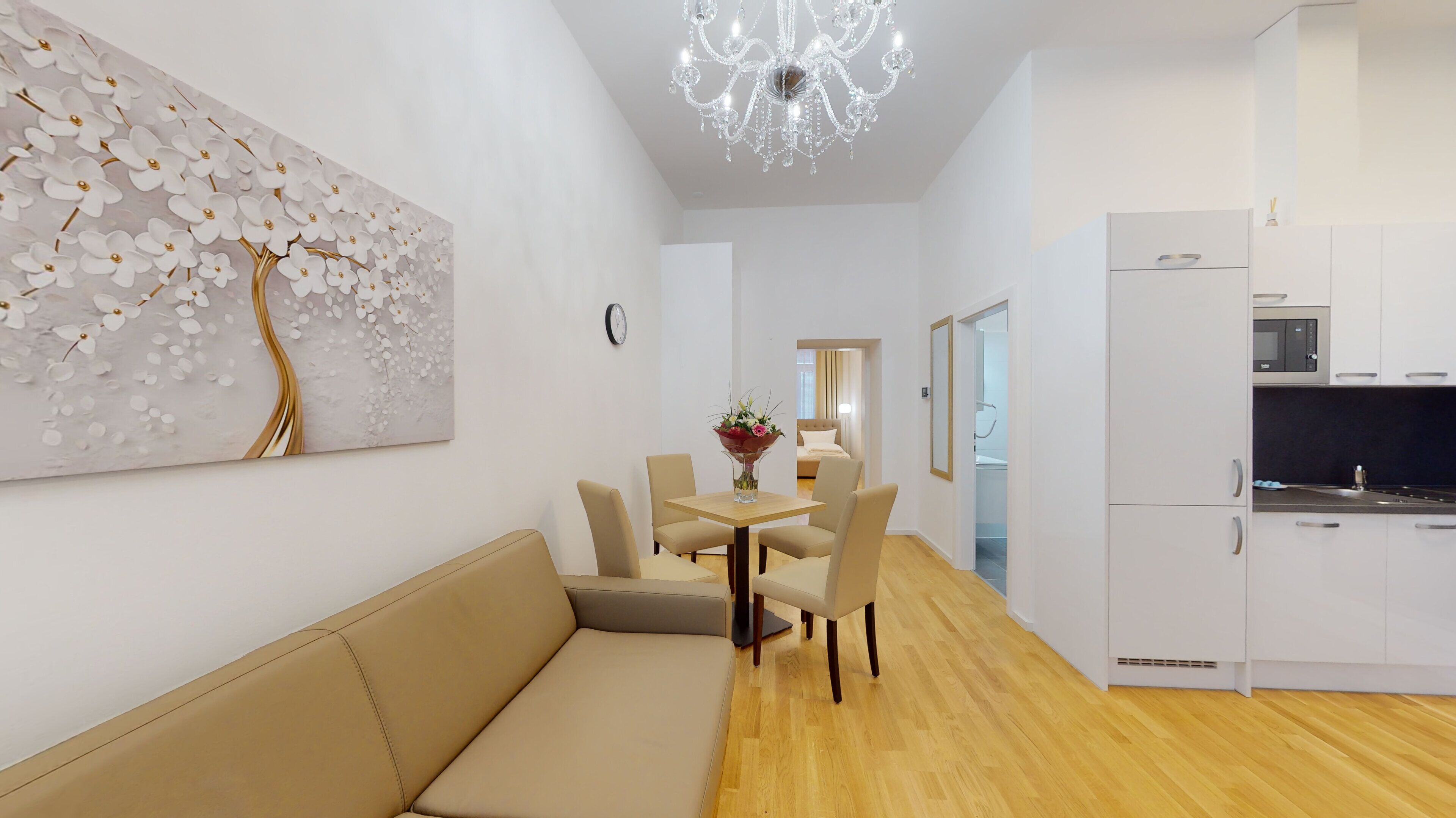 A modern living room with a beige sofa, a dining table with chairs, a chandelier, and a kitchen area with white cabinets and a microwave.