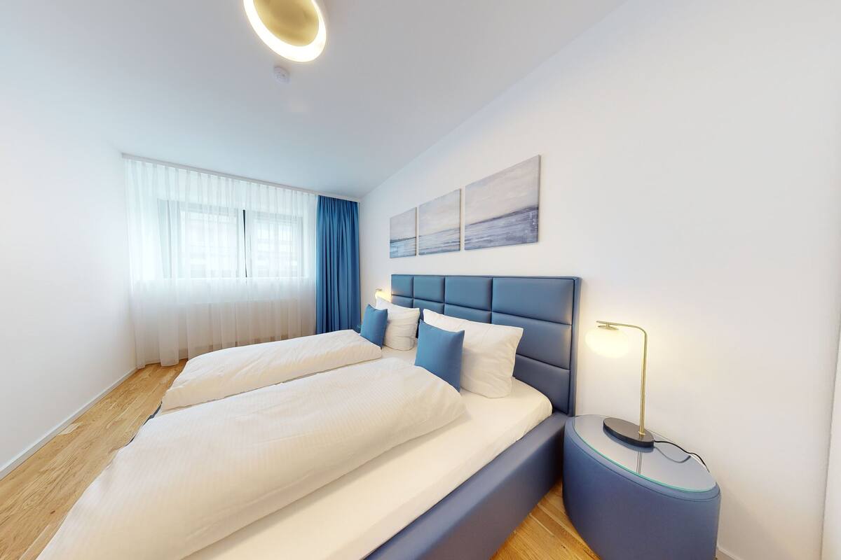 Une chambre d’hôtel moderne avec un grand lit, une table de chevet avec une lampe et une fenêtre ornée de rideaux légers.