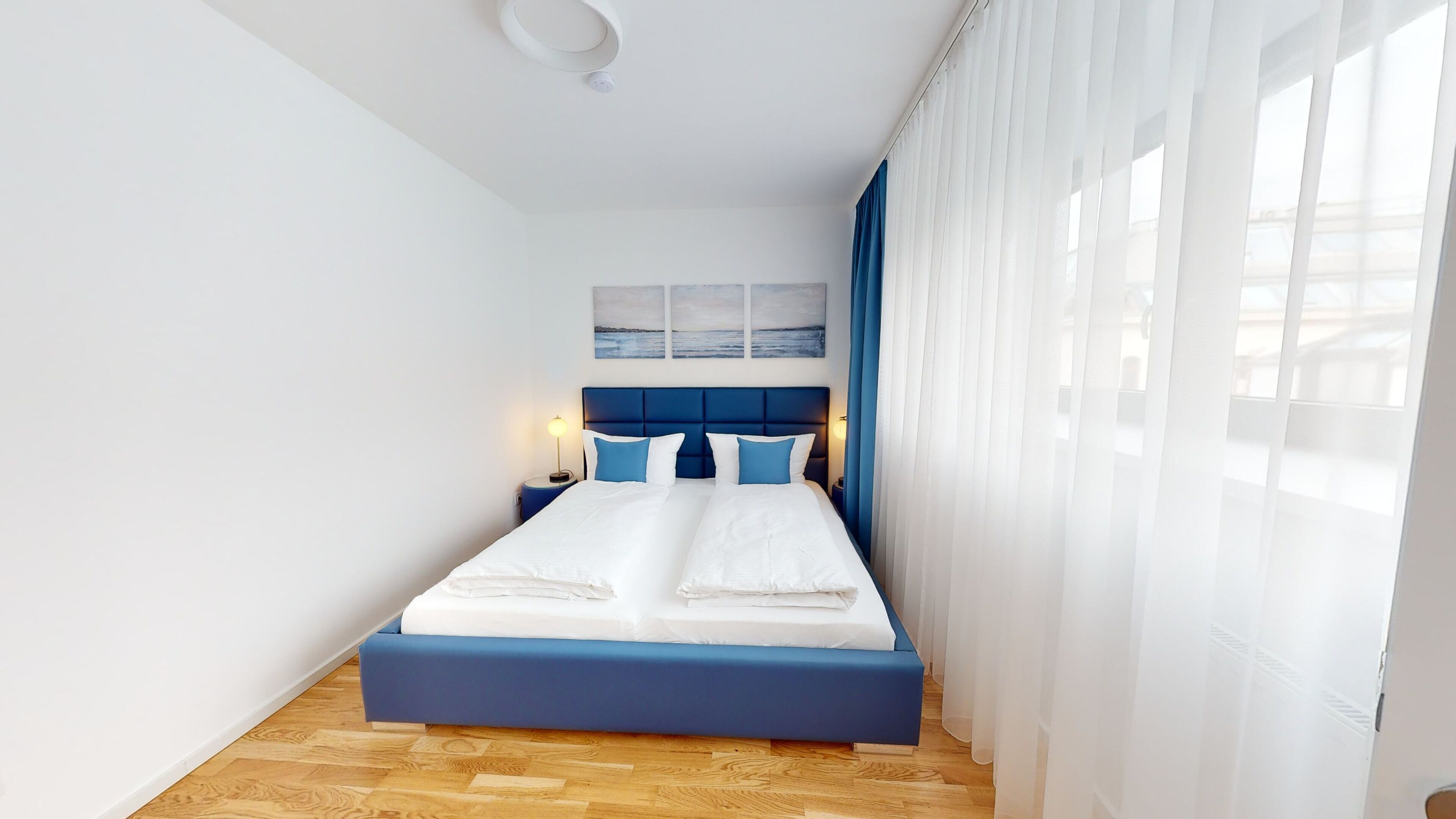 A modern hotel room with a large bed, a blue headboard, and a view of the city through sheer curtains.