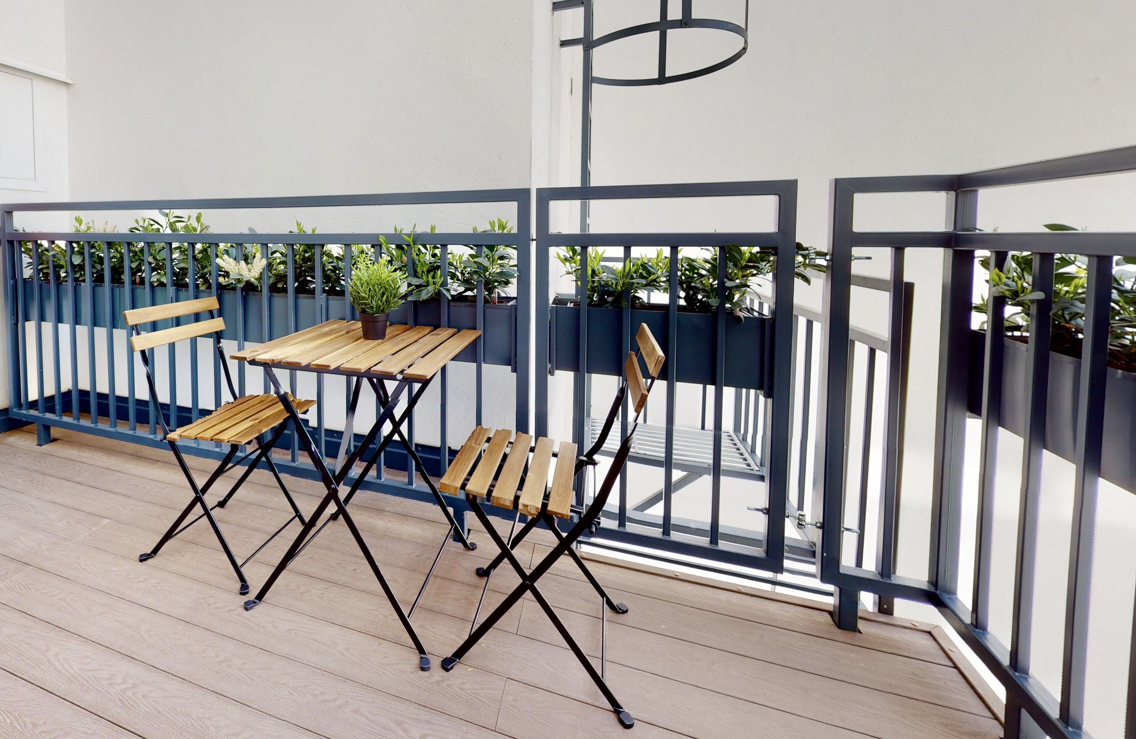A wooden deck with a table and two chairs, surrounded by a blue railing and potted plants.