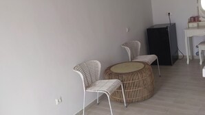 Una habitación con una silla blanca, un otoman de mimbre, una mesita y un armario negro.