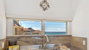 A kitchen with a stainless steel sink, a clock on the wall, and a window overlooking a coastal view. - Modern Condo W/Ocean Views and Lanai - Waikīkī Shore #ph07 (HONOLULU)