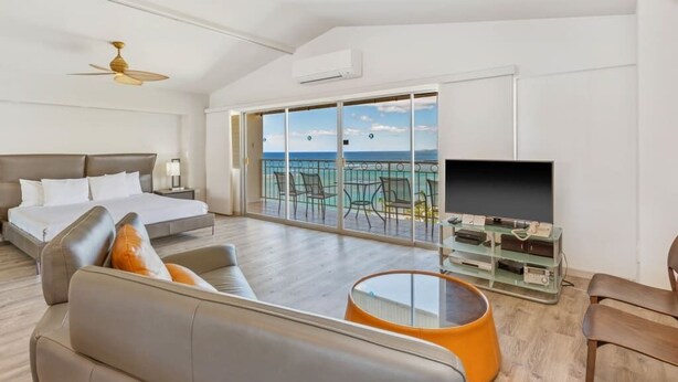 A modern hotel room with a balcony, a bed, a sofa, a TV, and a ceiling fan. - Modern Condo W/Ocean Views and Lanai - Waikīkī Shore #ph07 (HONOLULU)