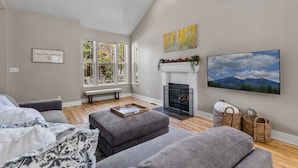 A modern living room with grey sofas, a fireplace, and a large window with a view of trees. - Modern Cabin Minutes to Flagstaff! Trailside Retreat w/ Trails/Park & 2 Car Garage (Flagstaff)