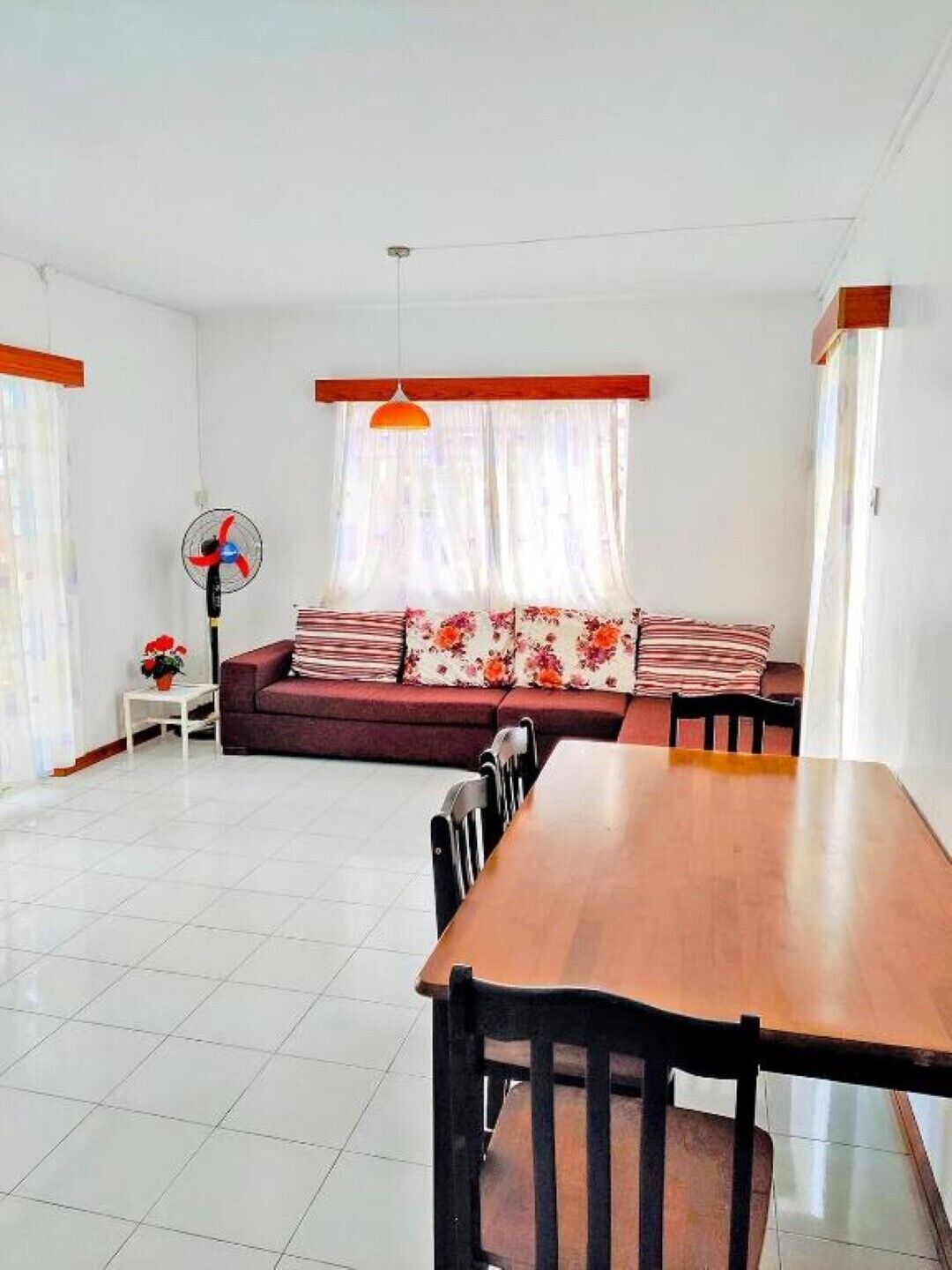 A modern living room with a wooden dining table, a sofa with floral cushions, a standing fan, and a window with curtains.