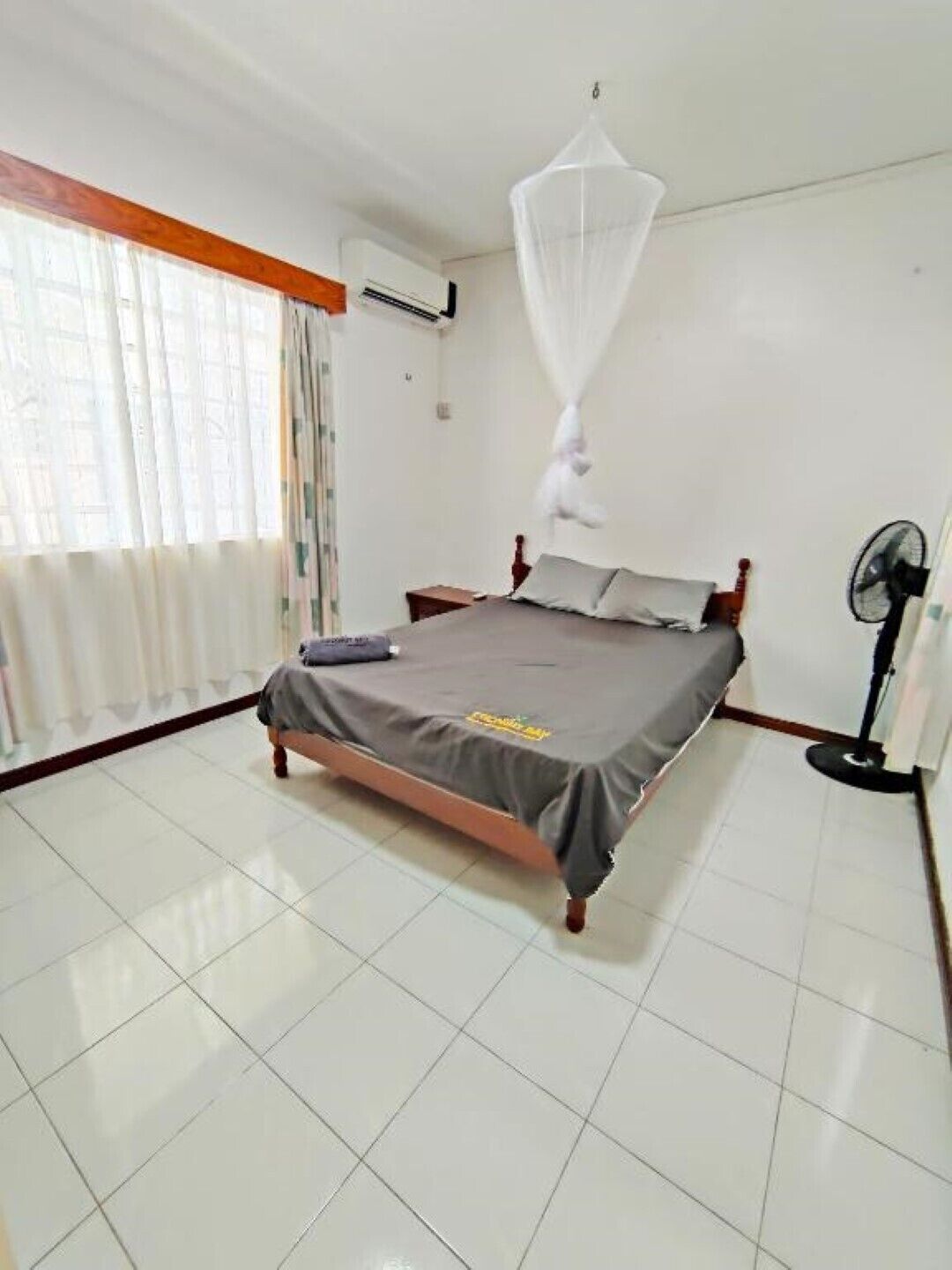 A bedroom with a bed, a standing fan, and a mosquito net.