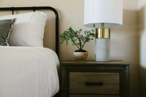A neatly made bed with white bedding and a single pillow, a bedside table with a lamp and a small potted plant, and a wooden nightstand with drawers.