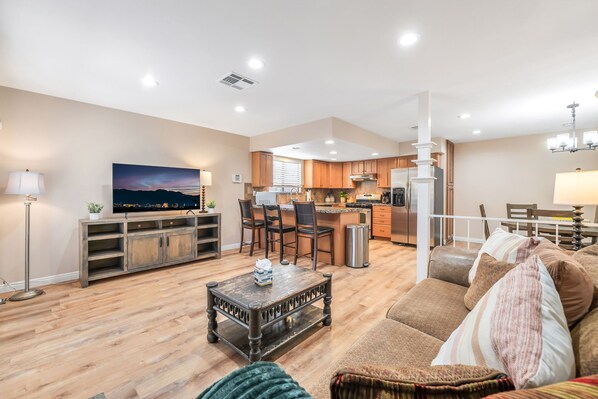 A modern living room with a sofa, coffee table, and a TV. There is a kitchen area with cabinets and a refrigerator.