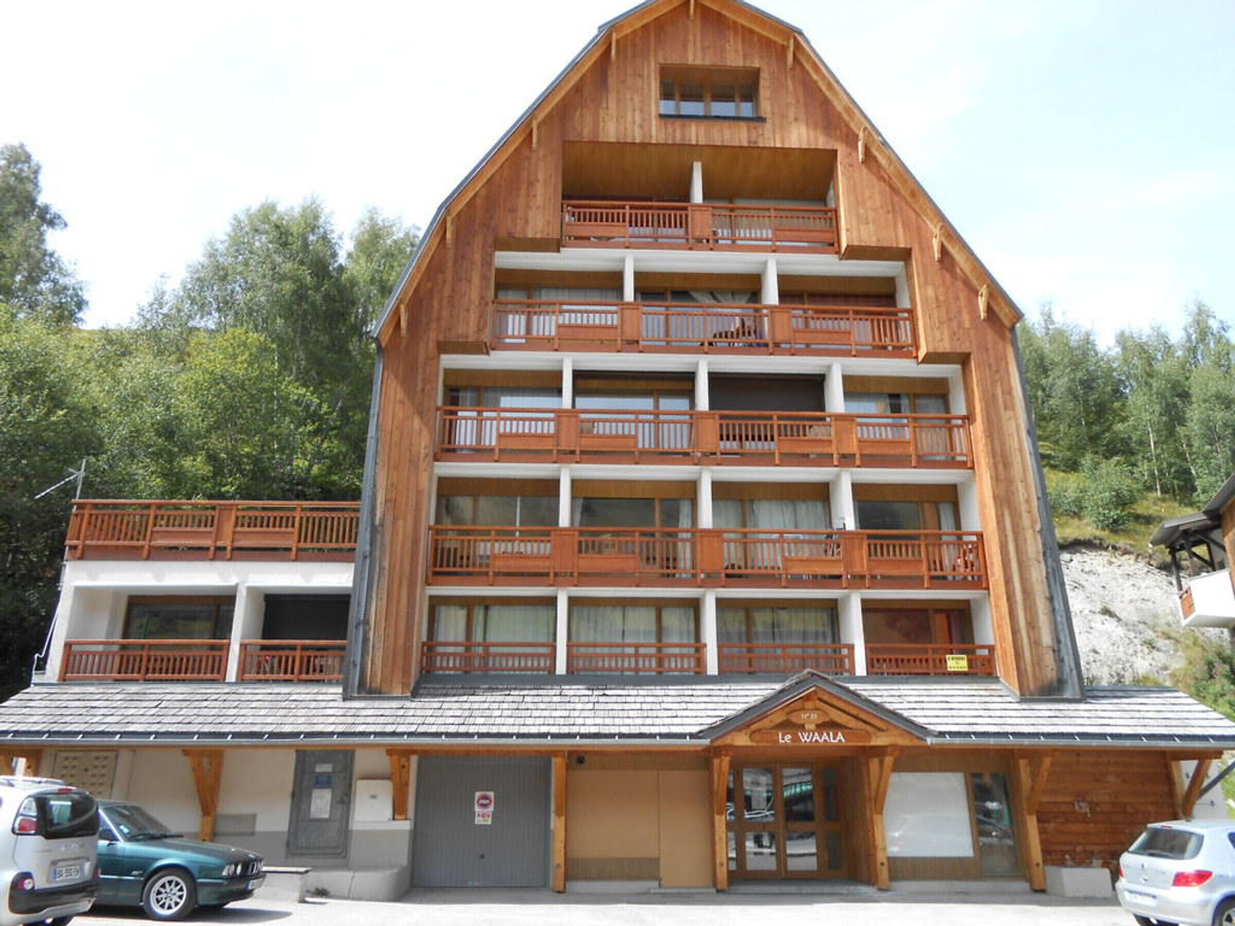 A multi-story wooden building with balconies, a sign reading "Le WAALA," and a parking area with cars.