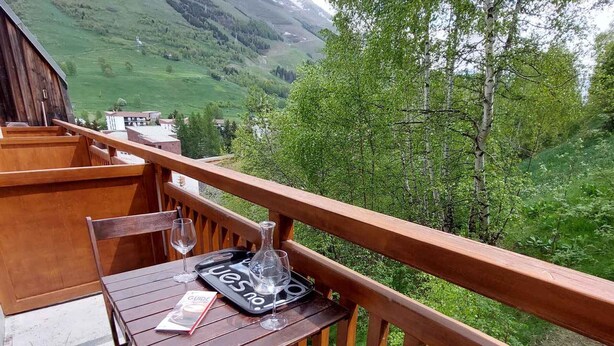 Une terrasse en bois avec une table et des chaises, un plateau avec des verres à vin et un guide touristique, donnant sur une vallée verdoyante.