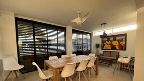 Un comedor con una mesa de madera y sillas blancas, ventanas amplias con persianas, un ventilador de techo y un cuadro en la pared.