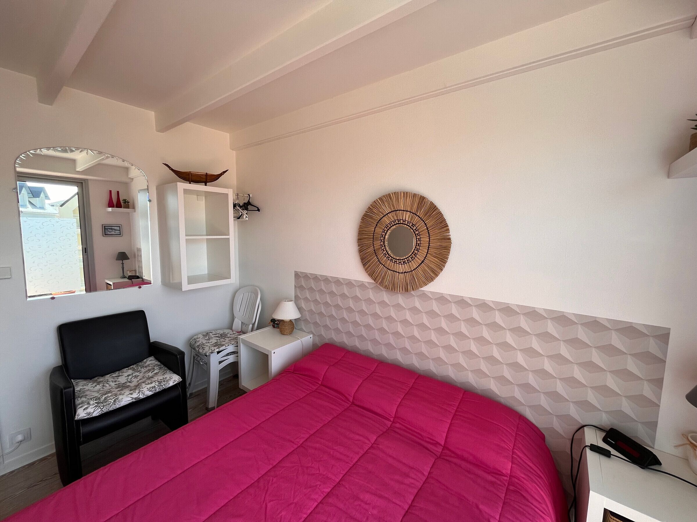 Een slaapkamer met een bed, een stoel, een klein tafeltje, een spiegel en een decoratief item aan de muur.