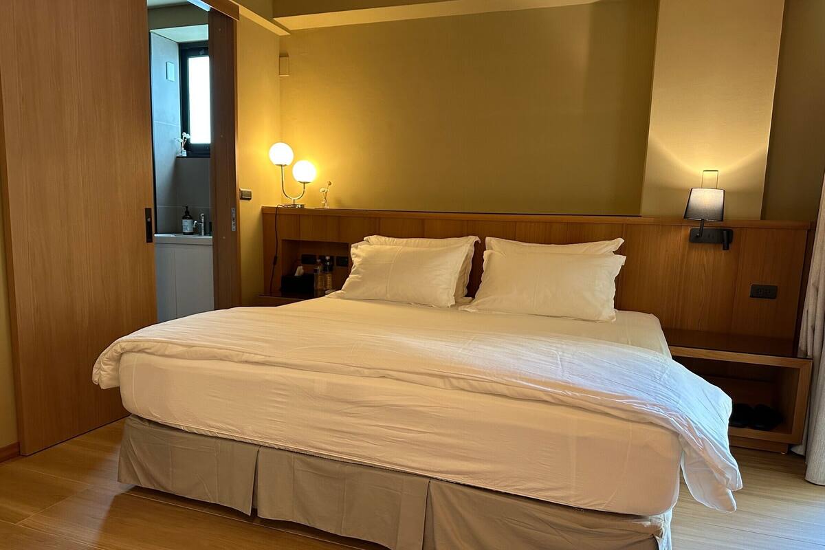 A neatly made bed with white linens, a wooden headboard, bedside lamps, and a small nightstand with a bottle.