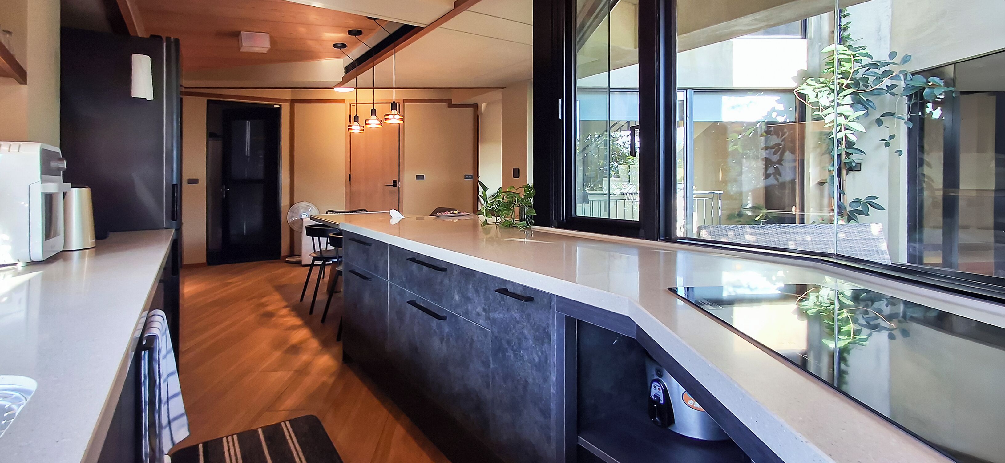 A modern kitchen with a central island, wooden flooring, and large windows offering a view of the outdoors.