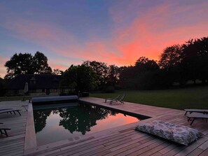 Una piscina con muelle de madera, sillas de descanso y una casa al fondo durante el atardecer.