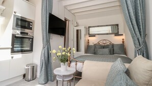Een knus slaapkamer met een bed, een klein tafeltje met bloemen, een televisie en een kitchenette met een oven.