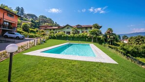 A swimming pool surrounded by a well-maintained lawn, with houses in the background and a clear blue sky. - Cottage del Lago: Lake Maggiore Villa With Shared Pool (Laveno Mombello)