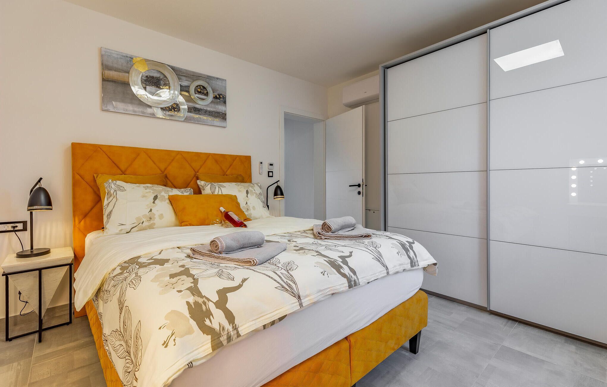 Un dormitorio moderno con un cabecero capitonado, una cama grande con ropa de cama floral, una mesita de noche con lámpara y un armario con puertas correderas.