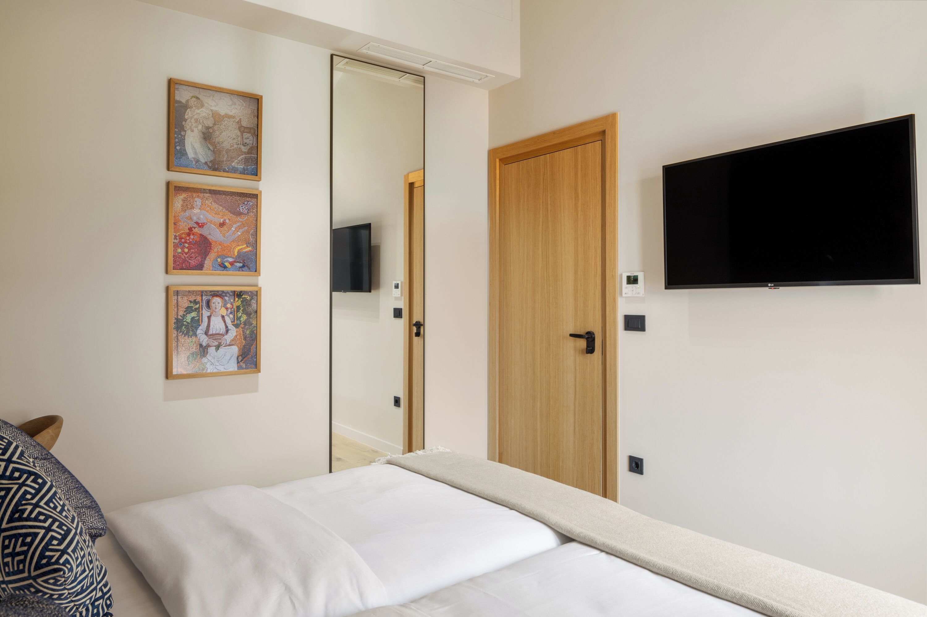 Quarto com cama, televisão presa na parede e três quadros enquadrados na parede.
