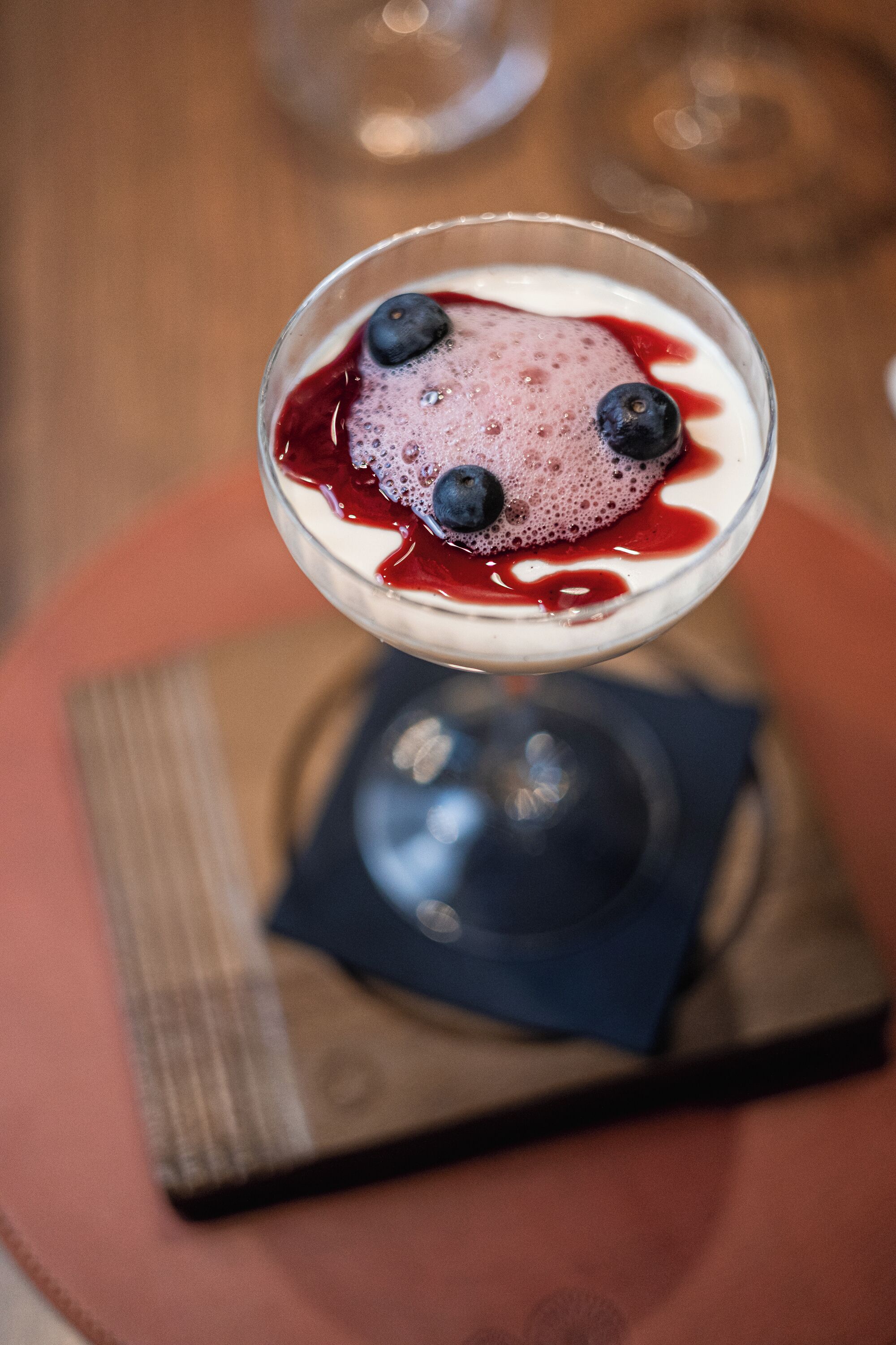Una copa de martini con una bebida cremosa, decorada con arándanos, sobre una mesa de madera con un mantel individual.