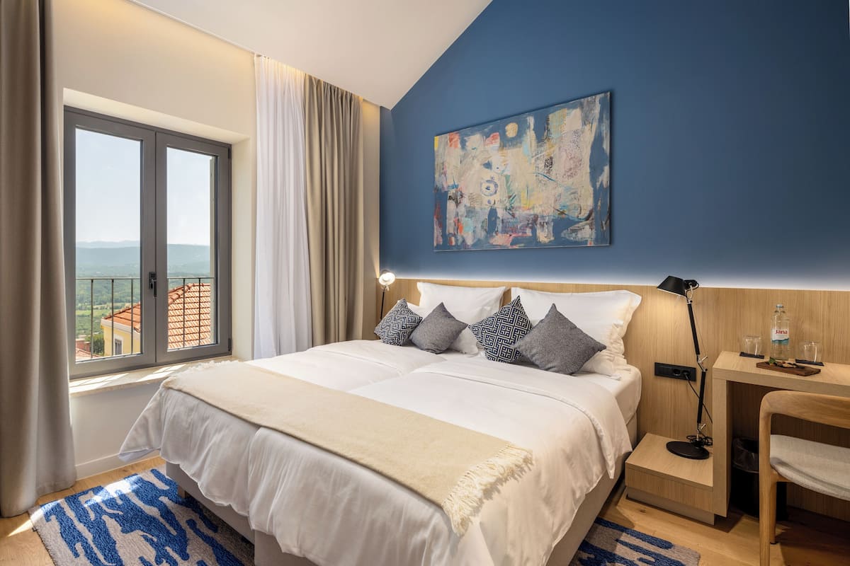 Une chambre avec un grand lit, une table de chevet, un bureau, une chaise, un tableau au mur et une vue par la fenêtre.