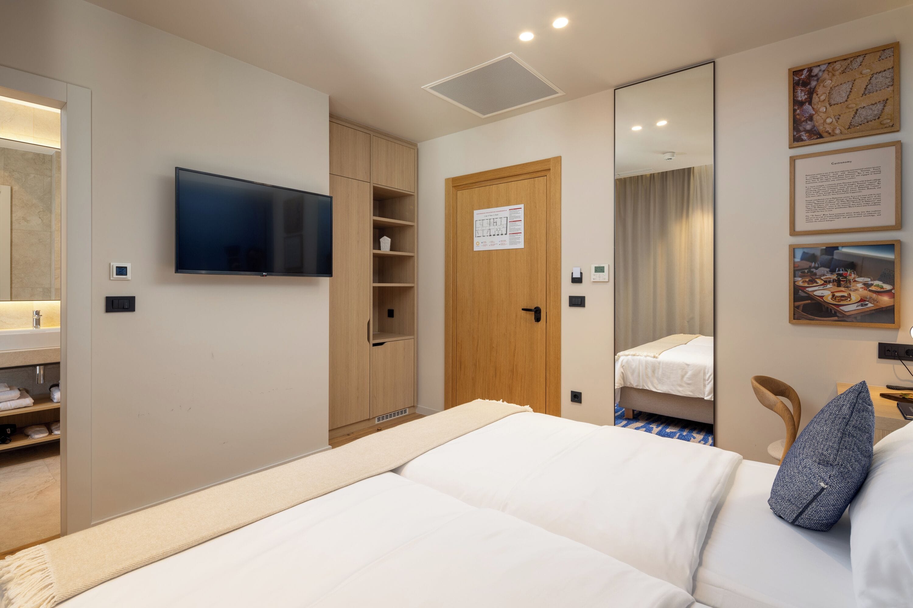 Una habitación de hotel con una cama, un televisor montado en la pared, un armario, un espejo y una mesita con una silla.