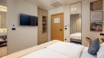 Una habitación de hotel con una cama, un televisor montado en la pared, un armario, un espejo y una mesita con una silla.