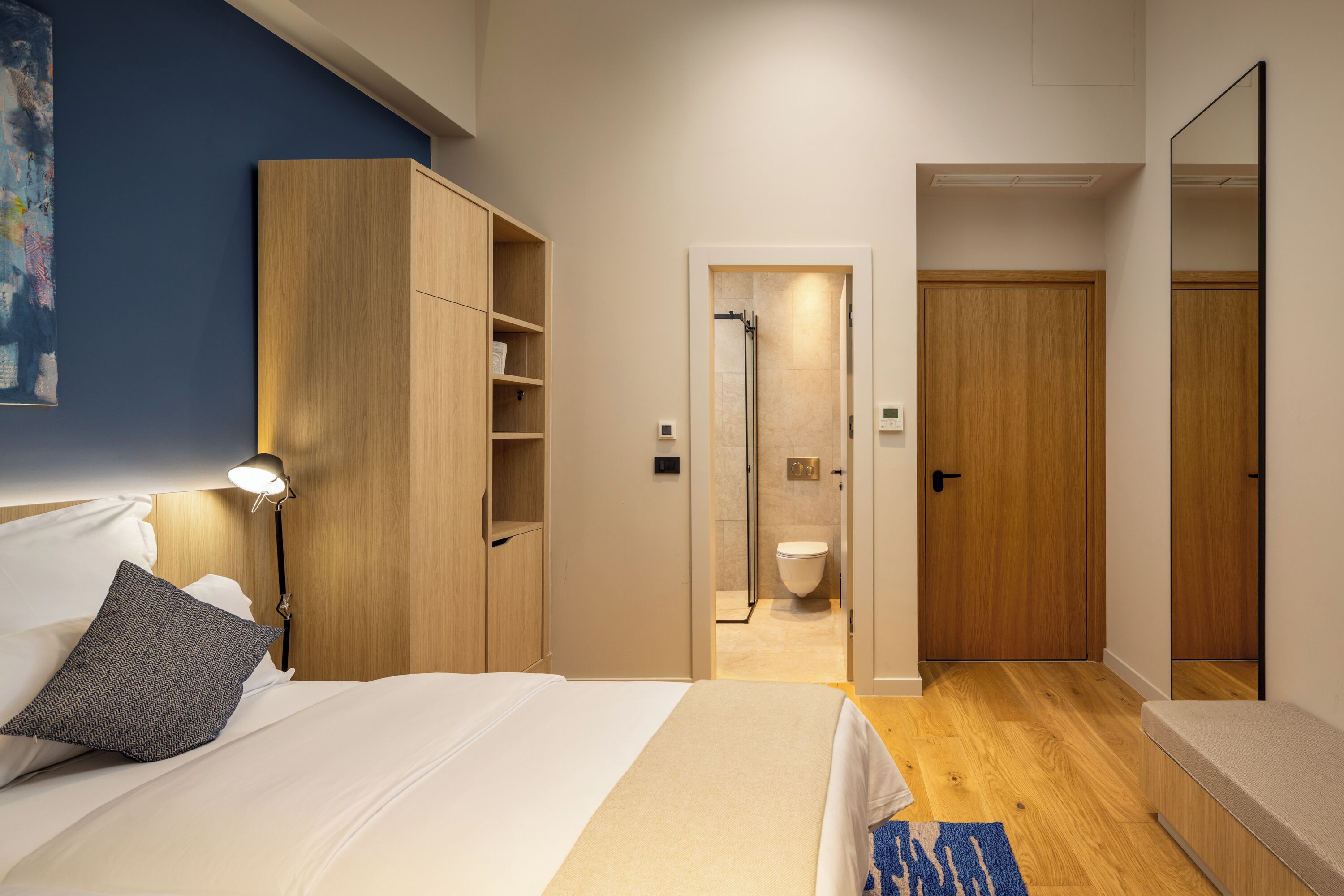 Ein modernes Hotelzimmer mit Bett, Nachttisch, Lampe, Schrank und einem durchgegangenen Tür zu sichtbarem Badezimmer.