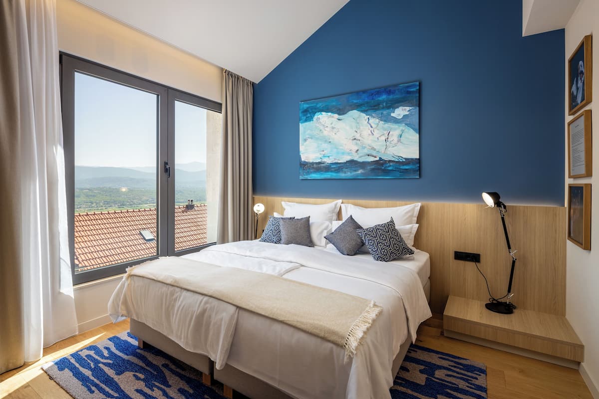 Une chambre avec un grand lit, une vue par une porte coulissante, un tableau au mur et une table de chevet avec une lampe.