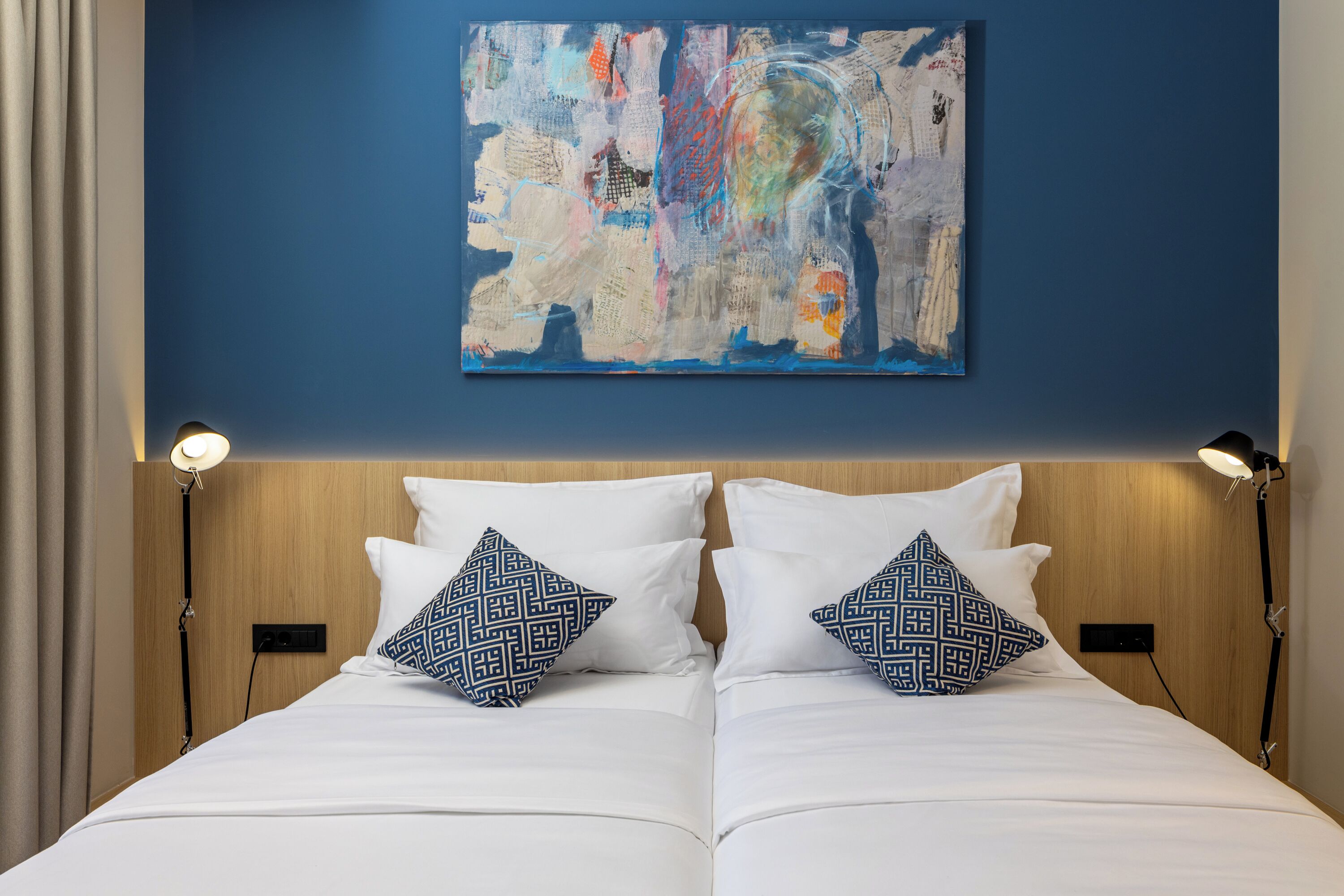 A double bed with white bedding and patterned pillows, a blue abstract painting on the wall, and two bedside lamps.