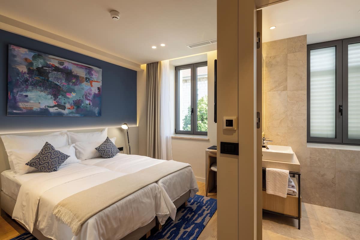Une chambre d’hôtel moderne avec un grand lit, un tableau au mur, une salle de bain avec un lavabo et un miroir, et une fenêtre avec des rideaux.