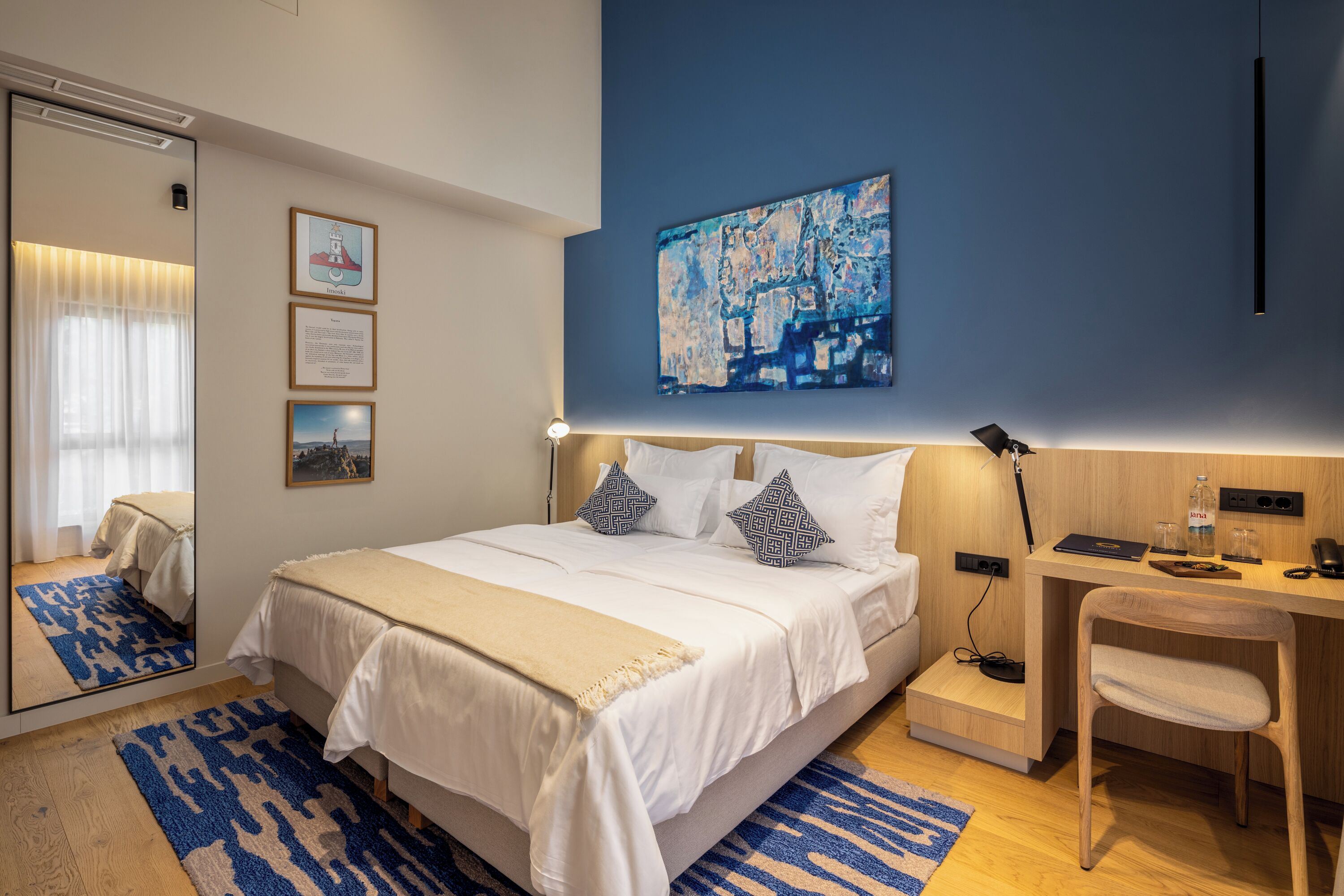 Et moderne hotelværelse med en stor seng, et skrivebord og en stol. Væggen er dekoreret med billeder i rammer, og der hænger et maleri over sengen.