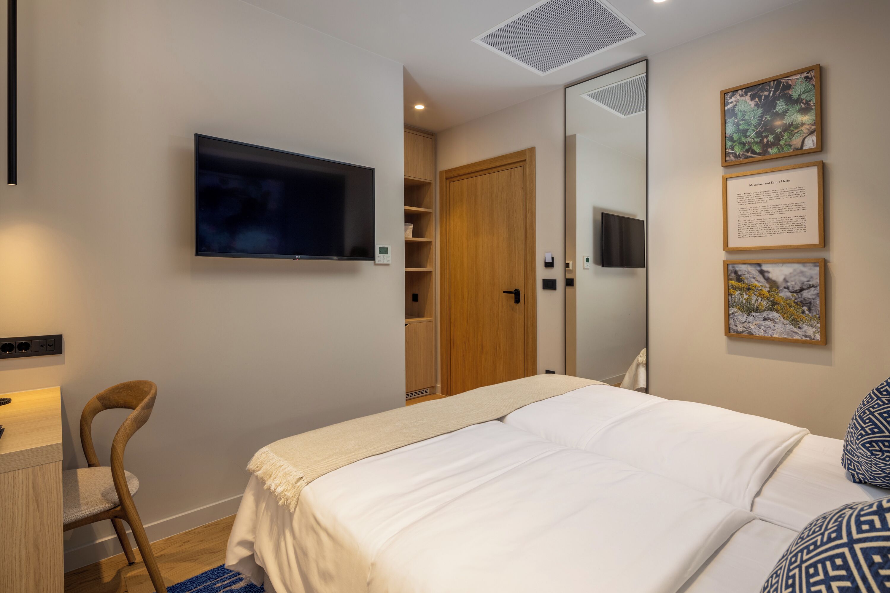 A bedroom with a bed, a TV mounted on the wall, a wooden wardrobe, and a desk with a chair.