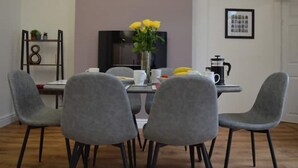 Zona pranzo con tavolo imbandito per quattro persone, sedie grigie, un vaso con fiori gialli, una macchina da caffè e un quadro appeso al muro.