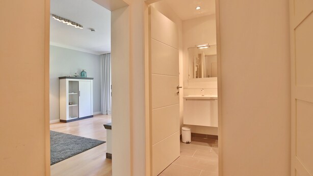 Una habitación de hotel moderna con suelo de madera, alfombra gris, armario blanco, una mesita con lámpara y un baño con espejo y lavabo.