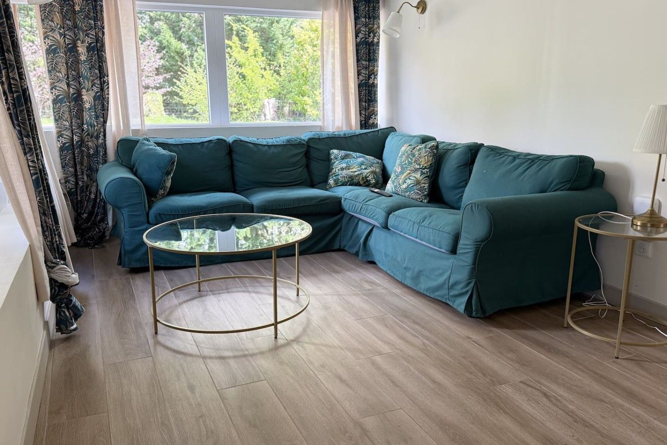 A corner living room with a teal sofa, a glass coffee table, and a lamp on a side table.